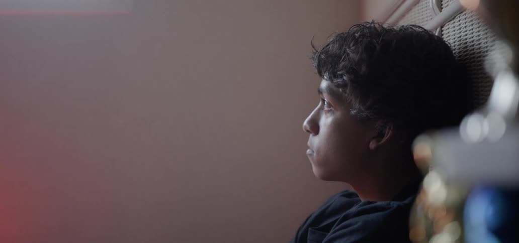 A boy is sitting on his bed in daylight thinking and staring up at a window