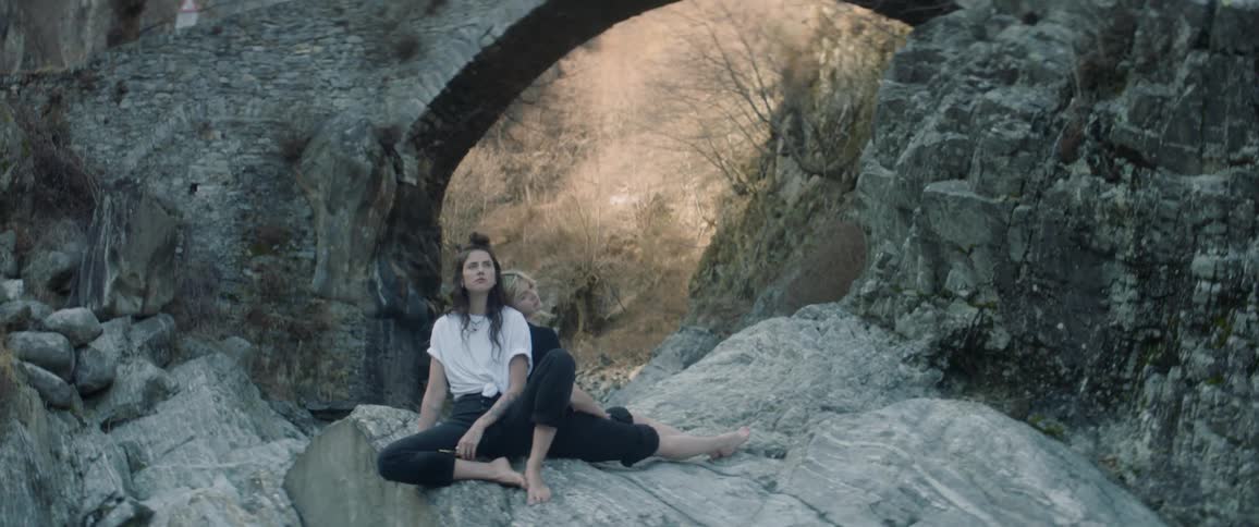 Two women lean against each other sitting on a large rock outside