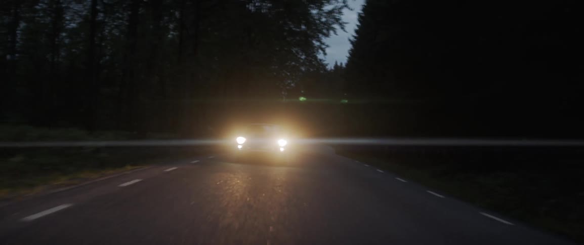 A car with headlights on travels on a tree lined rural road