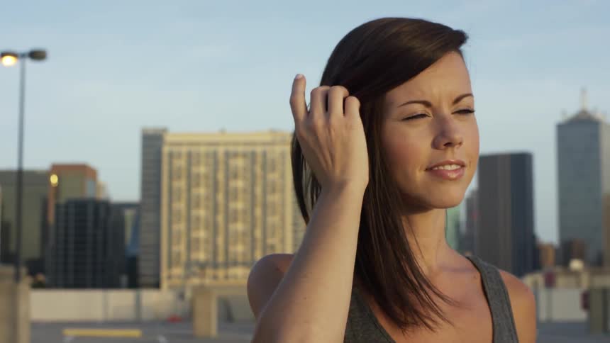 A brunette woman brushing her hair out of her face and looking off into the distance in an urban setting