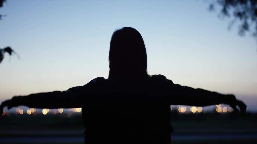 A woman turning towards lights that are off in the distance while raising her arms
