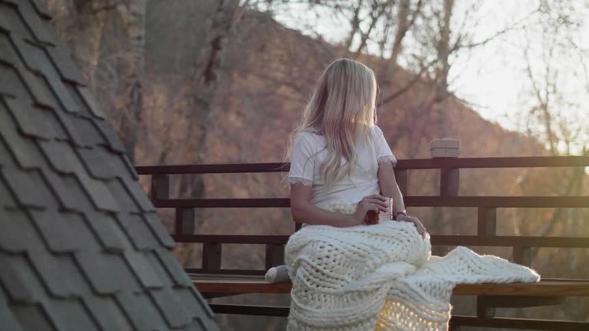 A woman sits wrapped in a blanket on a cabin balcony at sunset