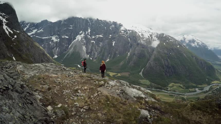 Two people hike in a rugged setting with snow capped mountains in the background