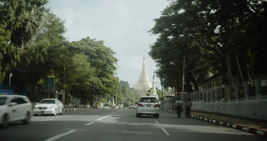 Vehicles and pedestrians drive down a main street towards a monument on a sunny day