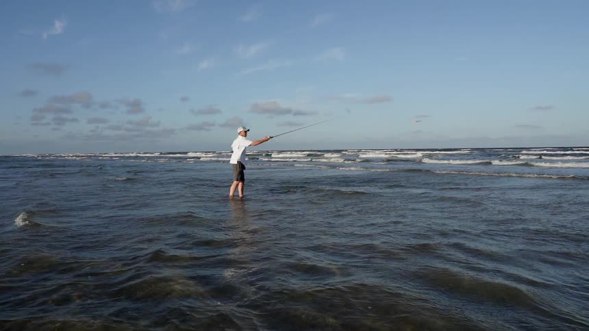 A man casts his fishing line into the ocean while standing in the surf