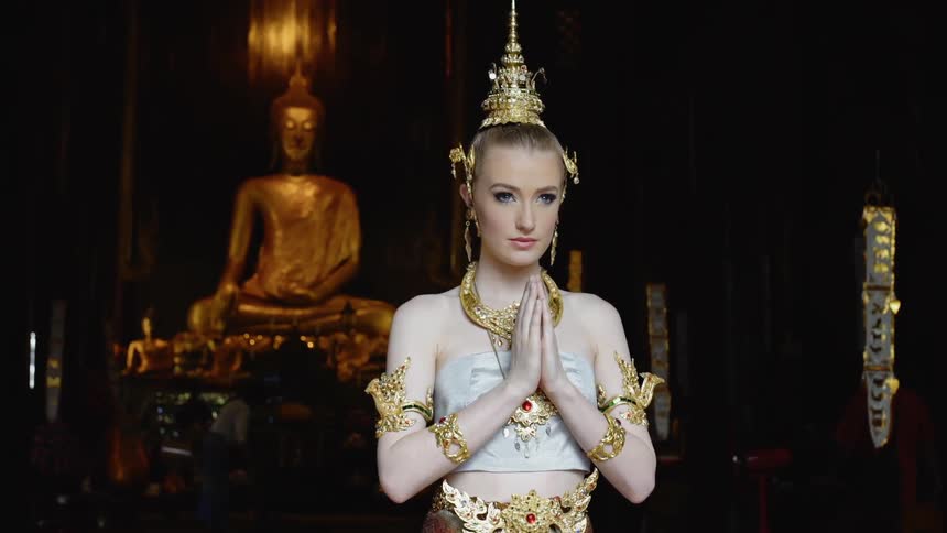 Woman wearing a headdress stands in a temple in front of a statue