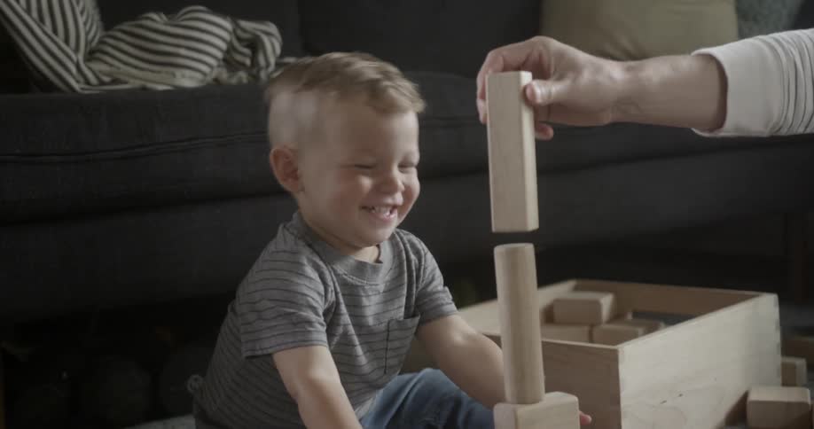 A toddler boy is laughing as he plays with wood bricks inside in daylight