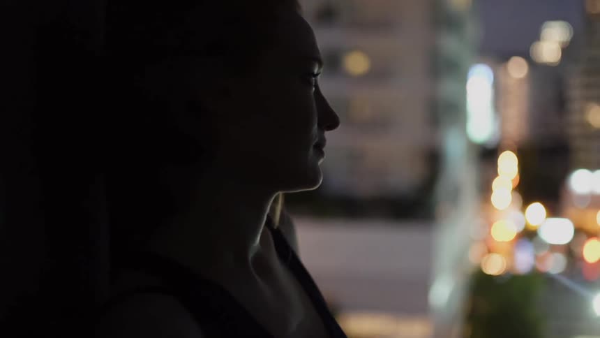 A woman stands on an apartment balcony in a city at night