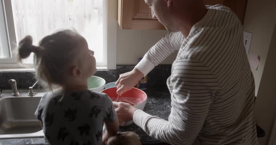 A dad stirs batter in a bowl while his children look on