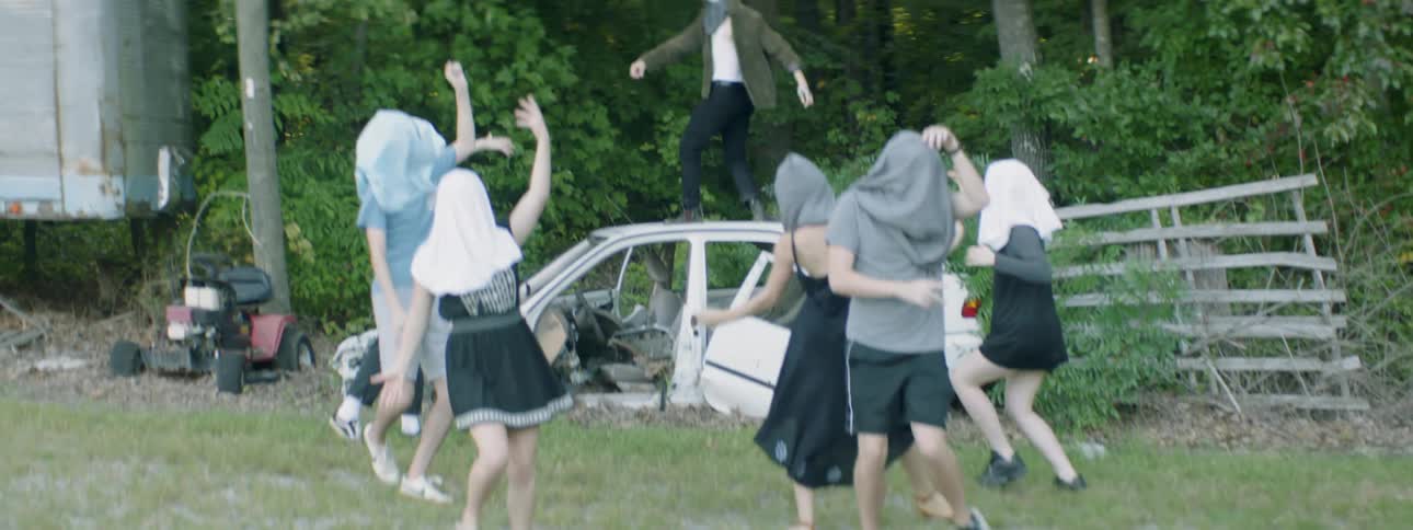 People with hoods over their heads dance around and on a wreck