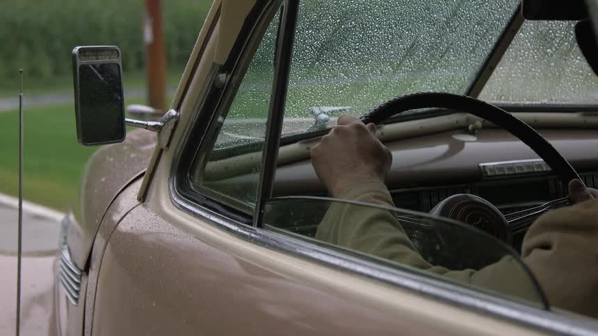 A man sits behind the wheel of an older model car on a rainy day