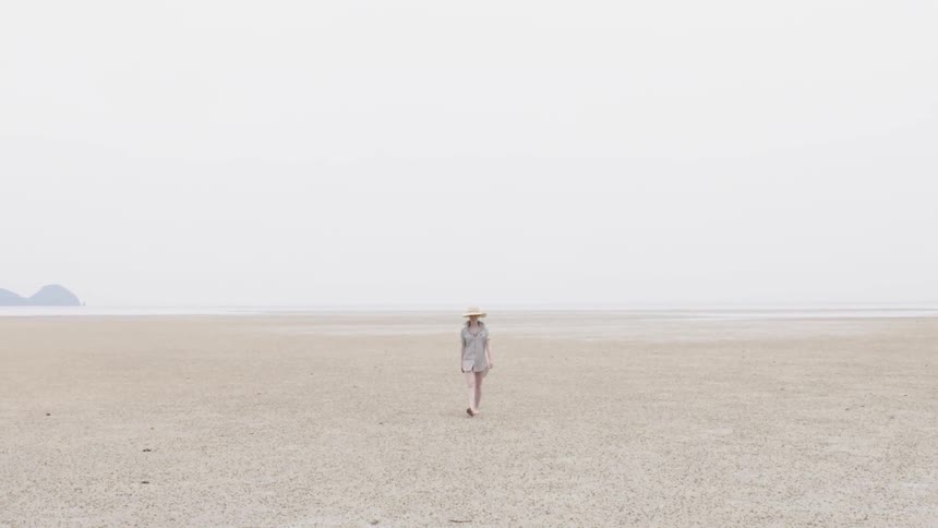 A woman wearing a hat walks on the sand away from the water
