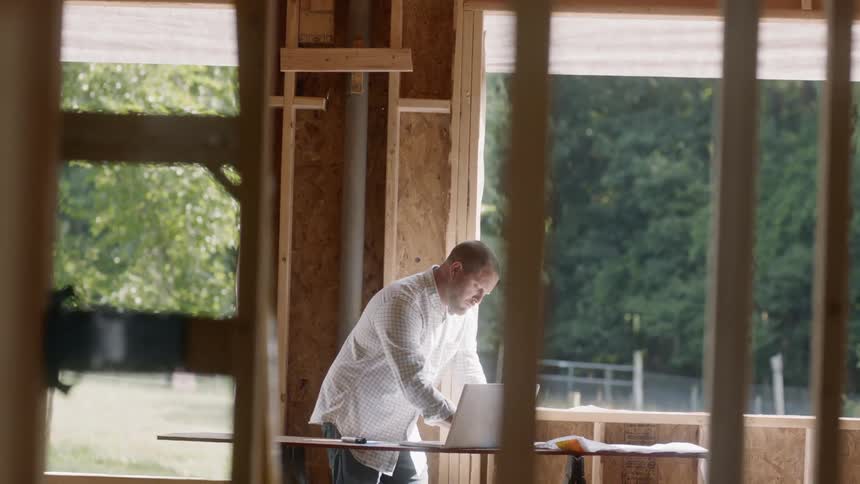 A man in a framed house works on a laptop