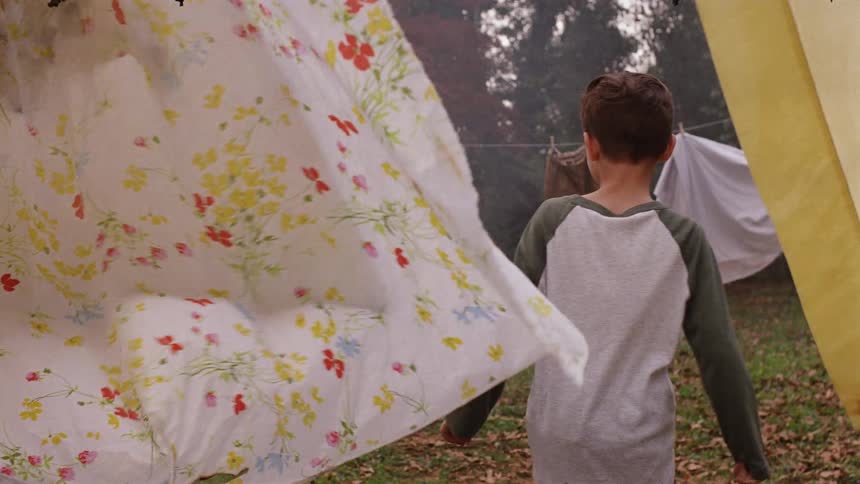 A woman is taking burned laundry off a clothesline as a boy sneaks up behind her in daylight