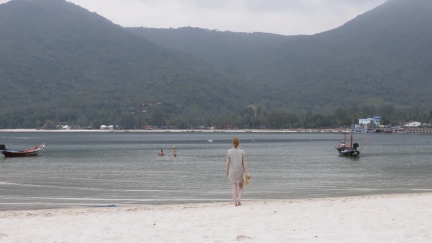 The woman removes her hat while walking towards the water