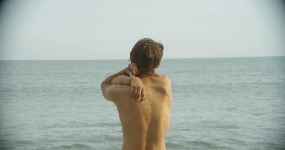 A shirtless man stretches his arms on a beach