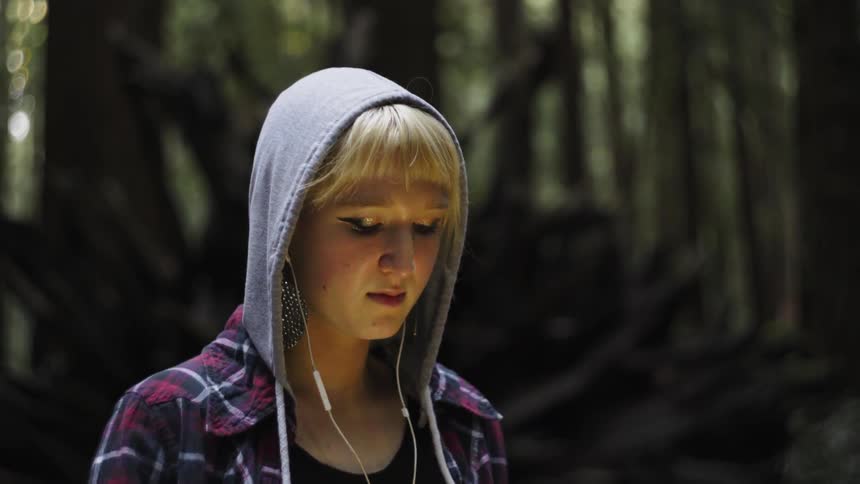 A teenage girl with a hoodie and earbuds on stands in a forest