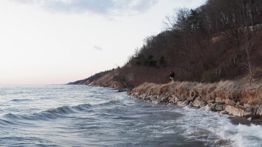 Two people are watching the waves crash against a beach wall on a cloudy day