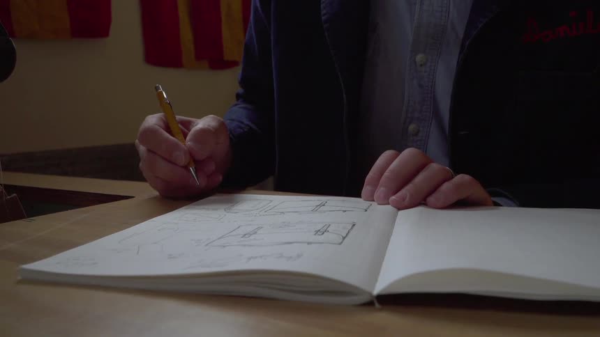 A man sits at a table holding a pencil and a notebook with sketches