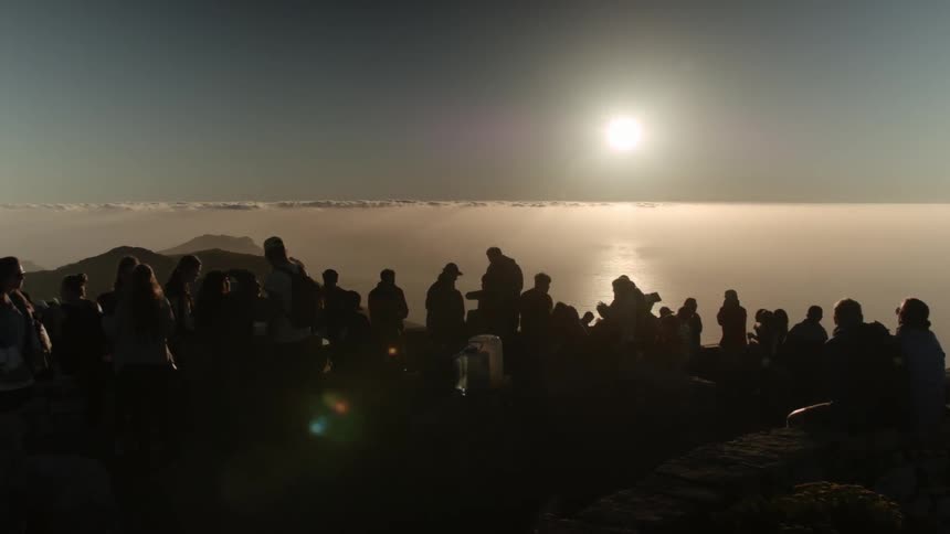 Numerous people gathered on top of a mountain overlooking other peaks and water as the sun sets