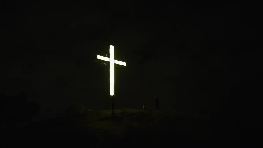 People walk towards and stand near a lit crucifix on a hill