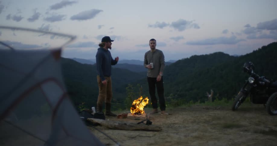 Two men talk outside of a tent by a fire in nature