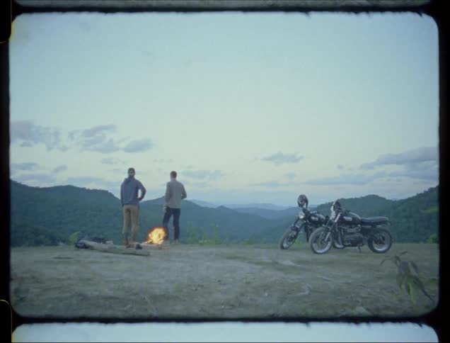 Two men stand atop a mountain as a fire burns