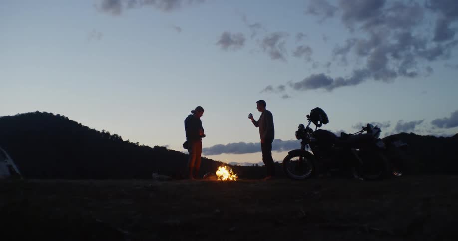 Two men are standing and drinking next to campfire at dusk