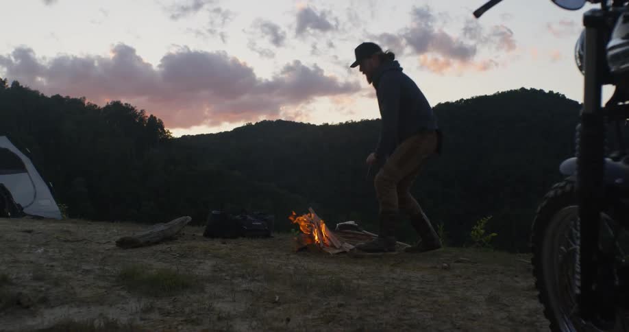 A man crouches next to a campfire at sunset