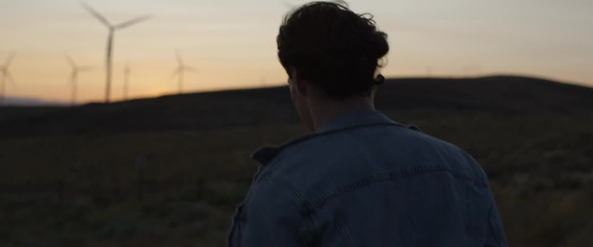 A man walks up a hill towards wind turbines at sunset