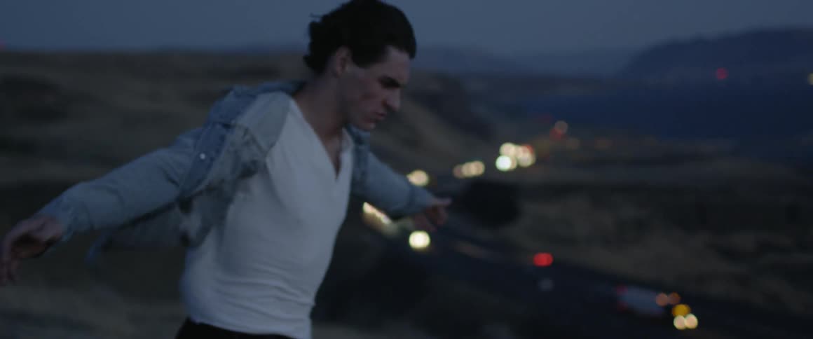 A man on a ridge leans into the wind with cars traveling below