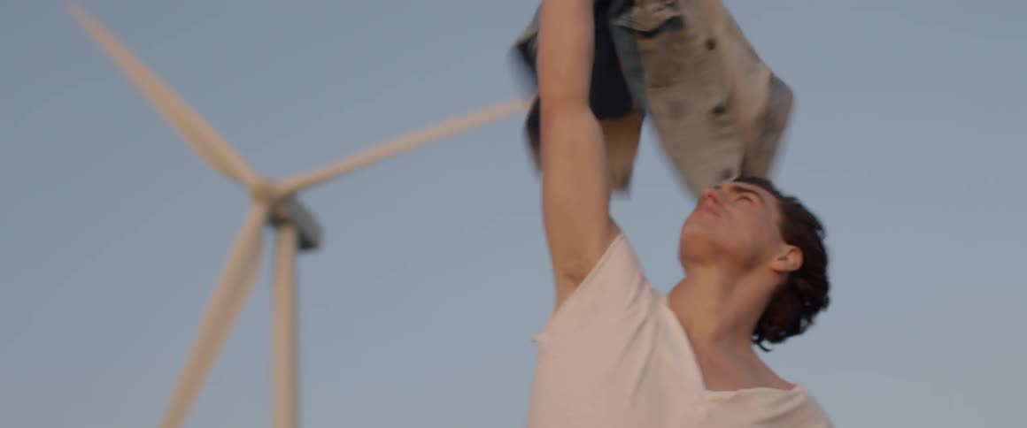 A man holds a denim jacket in the air near wind turbines