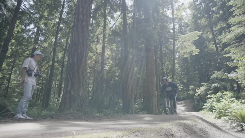 Man rides a bicycle over a path in the forest as someone with cameras watch