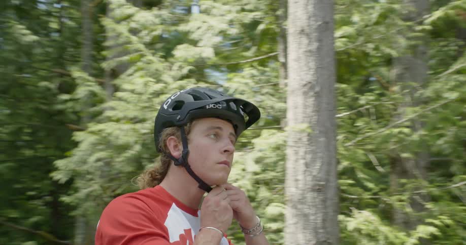 A man is putting on a helmet and riding off on his bike in a forest in daylight