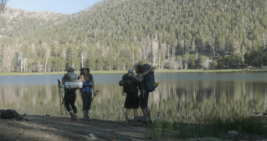 Four people wearing large backpacks greet each other at the edge of a lake