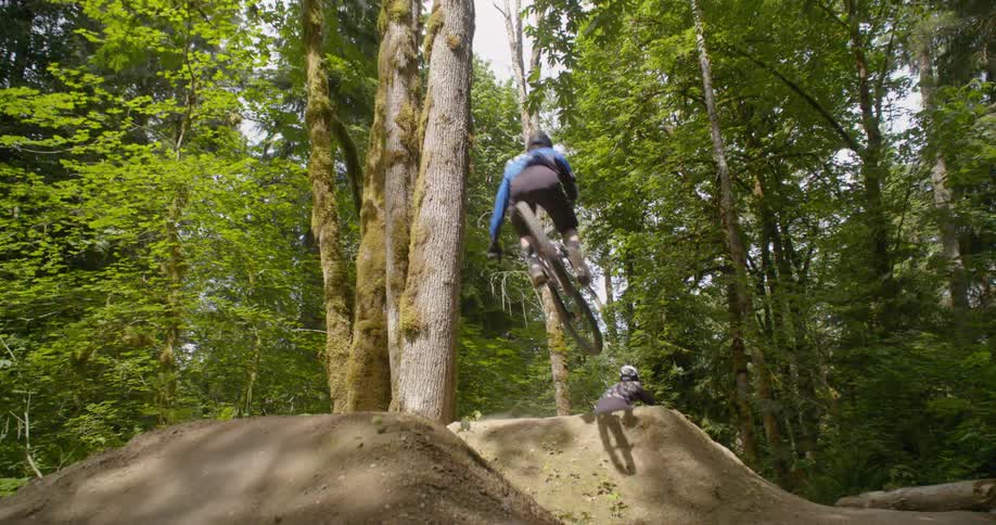 Mountain bikers ride over dirt ramps on a trail in the woods