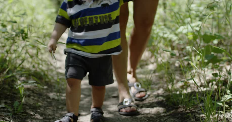 A mom holds her son's hand as they walk on dirt path in the woods