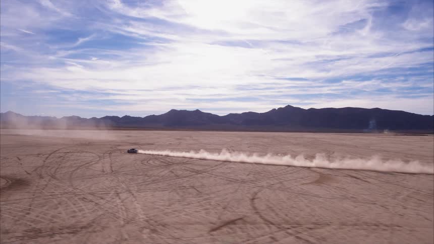 A car speeds across a dirt field surrounded by mountains