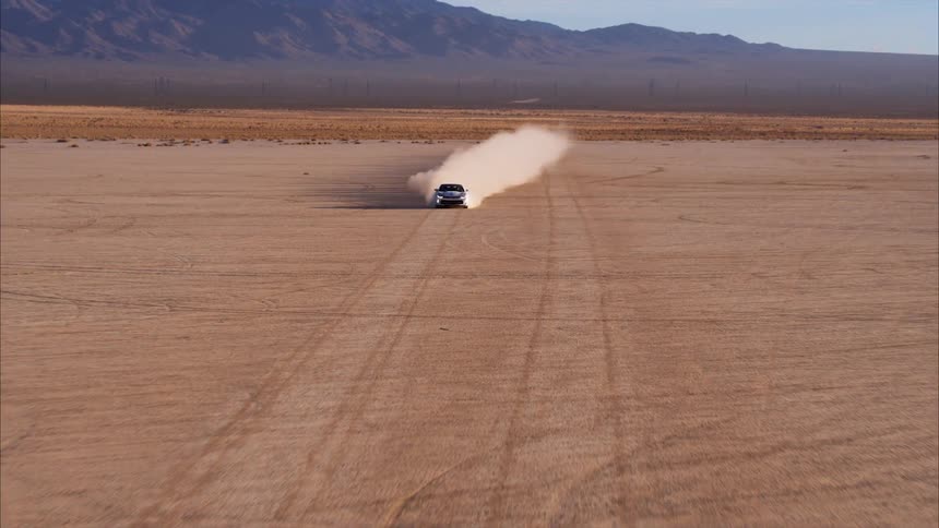 A car speeds across a dirt field surrounded by mountains