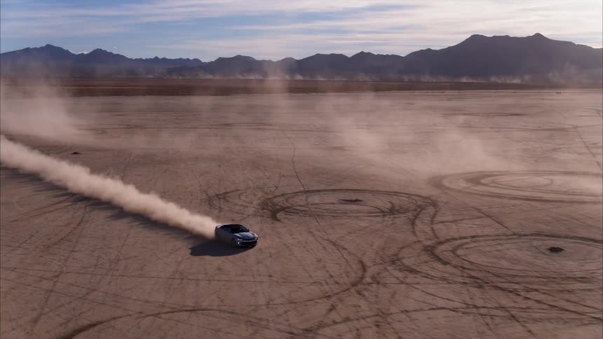 A car speeds across a dirt field surrounded by mountains
