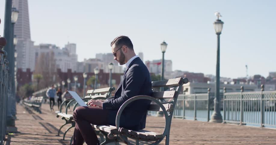 A man sits on a city bench using a laptop