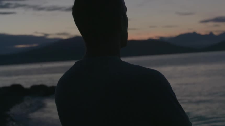 A man standing on the shoreline of a body of water looks out at a sunset