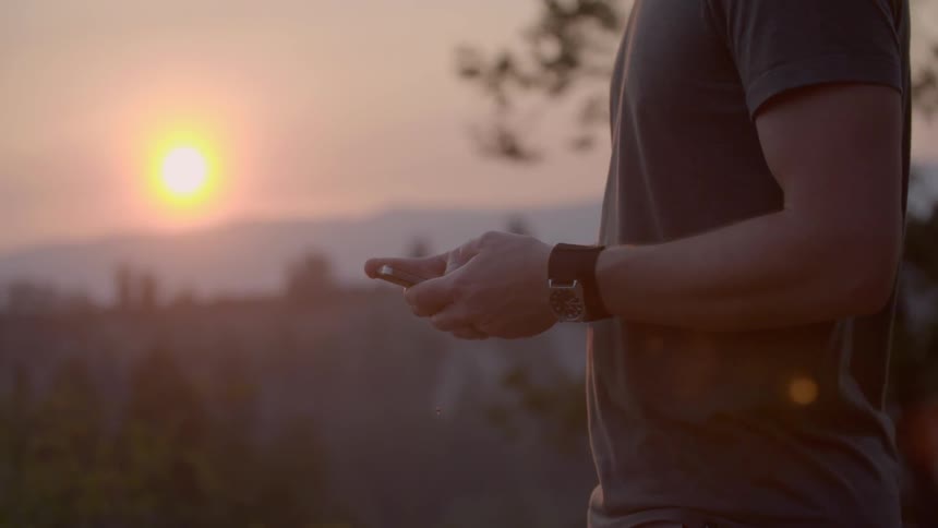 Man in natural setting fidgets with cell phone with sunset in the background