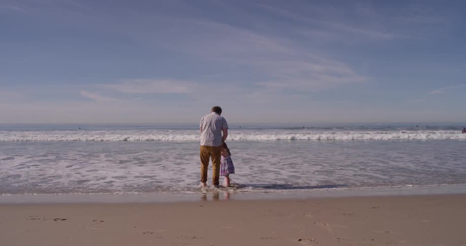 A man and young girl stand on the beach as the tide flows in
