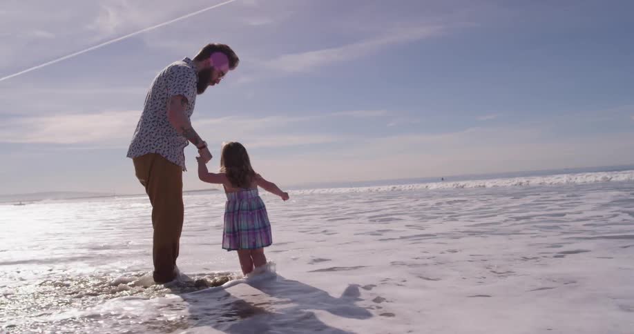Man and child stand on the beach and the water rolls over their feet