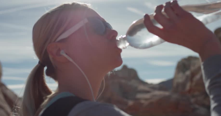 A woman drinks from a water bottle while wearing headphones in a desert