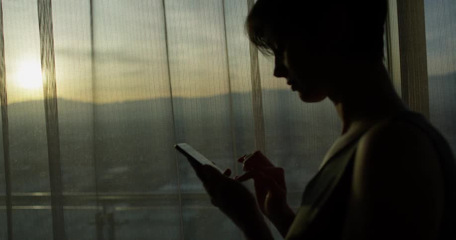 Woman stands in front of a large window at sunset looking at her phone