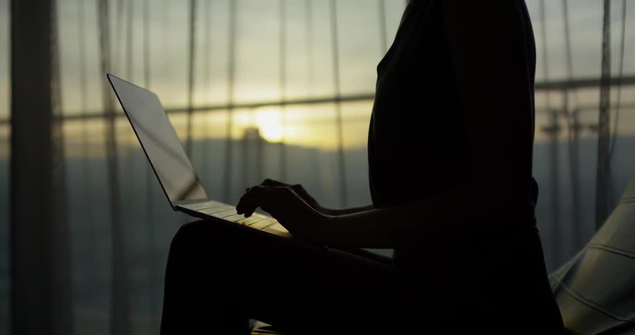 Person types on a laptop next to a large window at sunset