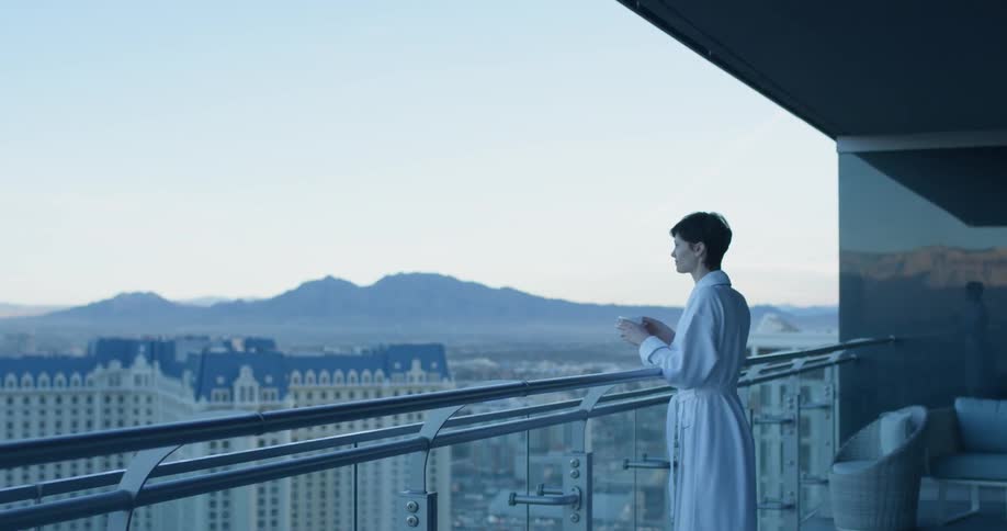 A woman has a drink looking over the city from the balcony