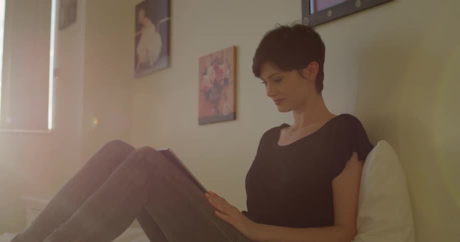 Woman sits on a bed and scrolls on a tablet